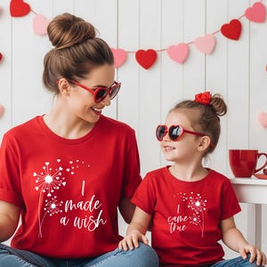 May include: Two red t-shirts with white dandelion graphics and text. One shirt says "I made a wish," the other says "I came true." Both shirts are worn with heart-shaped sunglasses. A Valentine's Day theme.