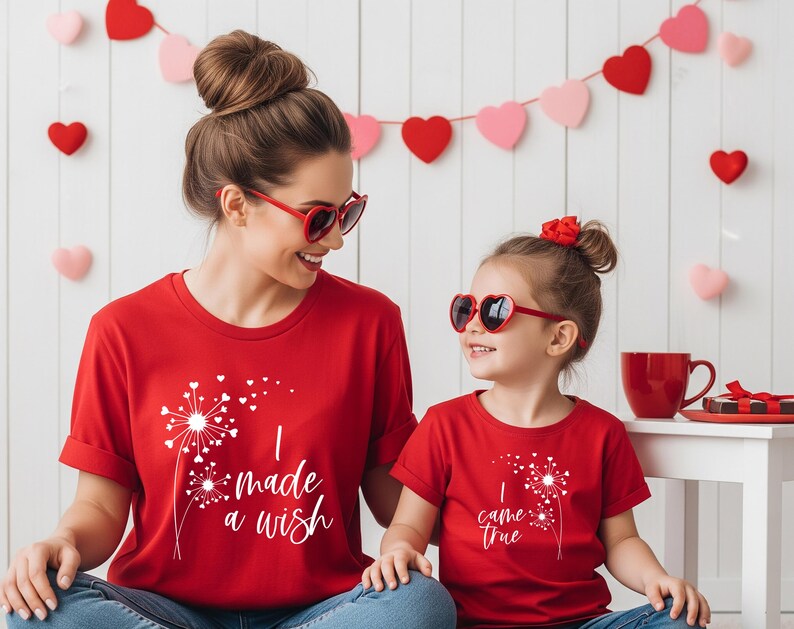 May include: Two red t-shirts with white dandelion graphics and text. One shirt says "I made a wish," the other says "I came true." Both shirts are worn with heart-shaped sunglasses. A Valentine's Day theme.