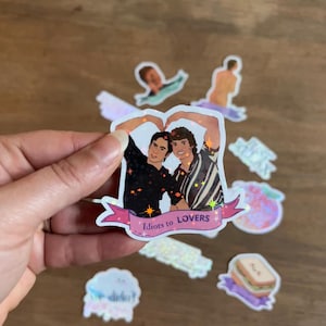 Puede incluir: Una pegatina con dos hombres formando un corazón con sus brazos. La pegatina tiene el texto "Idiots to LOVERS" en una pancarta rosa. Otras pegatinas holográficas están dispersas.