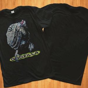 Può includere: Maglietta nera con una stampa grafica di Godzilla sul davanti. La grafica è un'illustrazione blu e verde della testa e del collo di Godzilla. Il testo "Godzilla" è stampato in verde sotto la grafica.