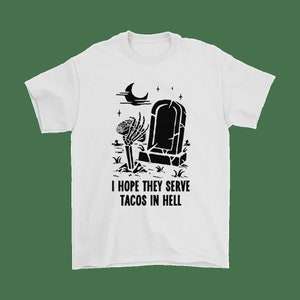 Pode incluir: Uma camiseta branca com um gráfico preto de um esqueleto segurando um taco na frente de uma lápide. O texto "Espero que sirvam tacos no inferno" está impresso abaixo do gráfico.