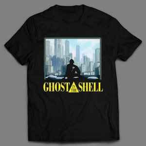 Può includere: Maglietta nera con una grafica gialla di una città e una persona seduta su un tetto. Il testo "Ghost in the Shell" è stampato in giallo sotto la grafica.