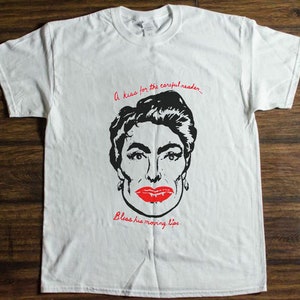 以下が含まれることがあります： 赤い唇をした女性の顔の白黒イラストが描かれた白いTシャツ。Tシャツには「A kiss for the careful reader... Bless his moving lips.」というテキストがプリントされています。