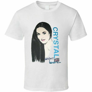 Może przedstawiać: Biała koszulka z niebiesko-białą ilustracją kobiety z długimi czarnymi włosami. Tekst "CRYSTAL GAYLE" jest nadrukowany pionowo po prawej stronie obrazu.