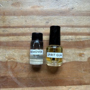 May include: Two small glass bottles with black caps. The left bottle contains a clear liquid and has a white label with the text REMOVER. The right bottle contains a yellowish liquid, has an applicator brush inside, and a white label with SPIRIT GUM.