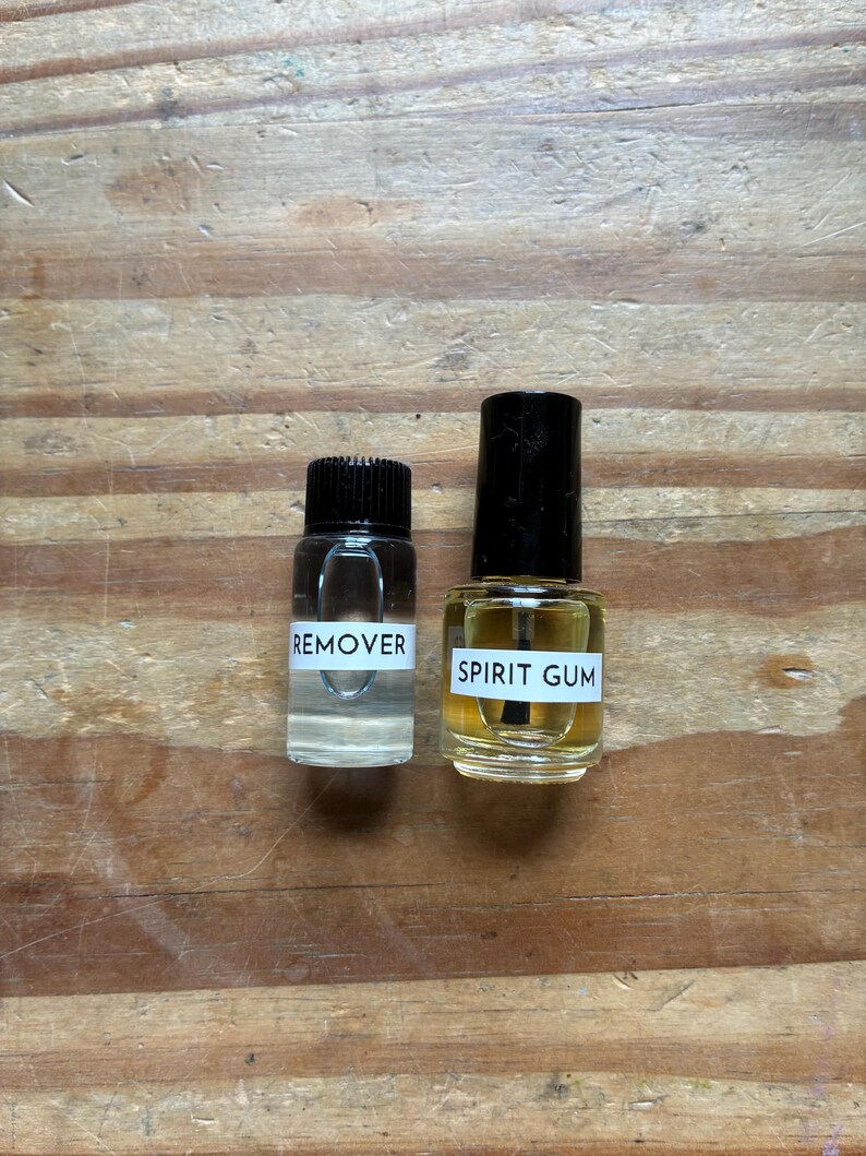 May include: Two small glass bottles with black caps. The left bottle contains a clear liquid and has a white label with the text REMOVER. The right bottle contains a yellowish liquid, has an applicator brush inside, and a white label with SPIRIT GUM.