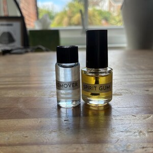 May include: Two small glass bottles with black screw-on caps. The left bottle contains a clear liquid and is labeled REMOVER. The right bottle contains a yellow-tinted liquid and is labeled SPIRIT GUM, featuring an applicator brush inside.