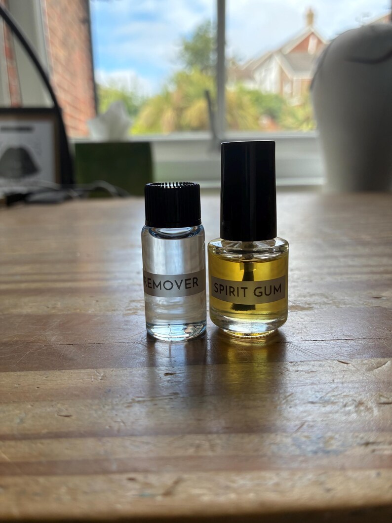 May include: Two small glass bottles with black screw-on caps. The left bottle contains a clear liquid and is labeled REMOVER. The right bottle contains a yellow-tinted liquid and is labeled SPIRIT GUM, featuring an applicator brush inside.