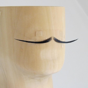 May include: A light-coloured wooden sculpture with a painted black moustache. The sculpture has a cylindrical shape with a rounded lower portion. The moustache is painted on the front of the sculpture.