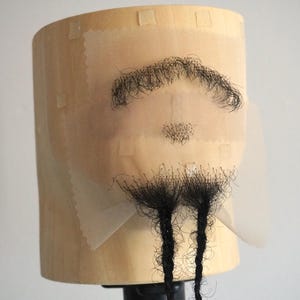Puede incluir: Un conjunto de vello facial teatral que incluye un bigote negro, una pequeña perilla y una barba larga con dos mechones trenzados. El pelo está sujeto a una base de encaje beige transparente, montada sobre un maniquí de madera de color claro.