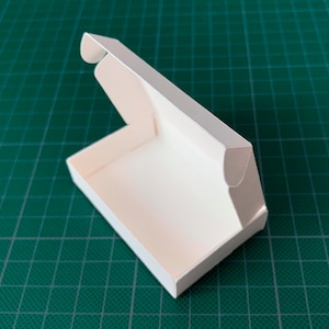 May include: A white cardboard box with a hinged lid, open to reveal the empty interior. The box is sitting on a green surface.