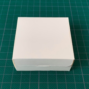 May include: A plain white cardboard box with a folded top flap. The box is sitting on a green surface.