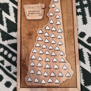 May include: Wooden wall art featuring a map of New Hampshire with 48 mountain peaks labeled. The sign reads "New Hampshire 48 4000 Footers." The artwork is framed in a light wood.