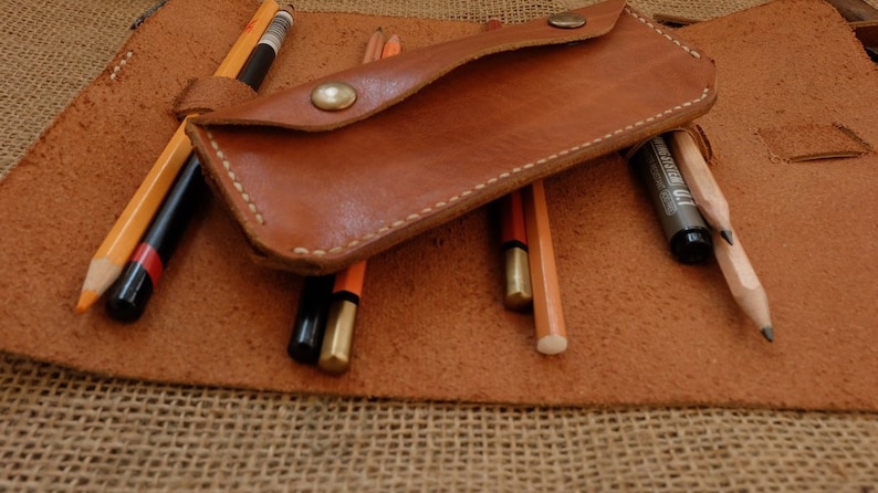 Puede incluir: Un estuche de l&aacute;pices de cuero marr&oacute;n con una solapa asegurada por un bot&oacute;n de lat&oacute;n. El estuche est&aacute; abierto, revelando varios l&aacute;pices de varios colores y dise&ntilde;os. Los l&aacute;pices descansan sobre una superficie de cuero m&aacute;s grande y abierta.