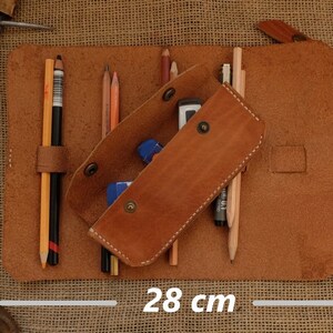 Puede incluir: Estuche de l&aacute;pices de cuero marr&oacute;n, de 28 cm de ancho y 20 cm de alto, con l&aacute;pices, gomas de borrar y un bol&iacute;grafo. El estuche est&aacute; abierto, revelando el contenido. El cuero tiene un aspecto natural y texturizado.