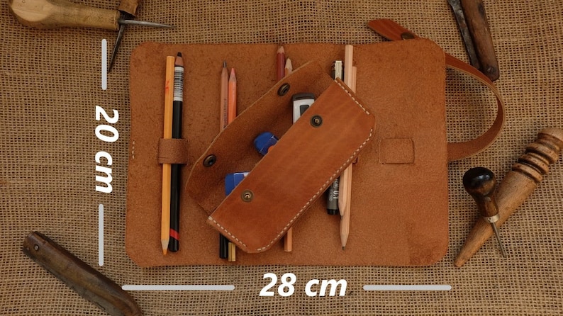 Puede incluir: Estuche de l&aacute;pices de cuero marr&oacute;n, de 28 cm de ancho y 20 cm de alto, con l&aacute;pices, gomas de borrar y un bol&iacute;grafo. El estuche est&aacute; abierto, revelando el contenido. El cuero tiene un aspecto natural y texturizado.