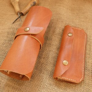 Puede incluir: Dos rollos de herramientas de cuero marr&oacute;n y un peque&ntilde;o estuche de cuero con cierres de bot&oacute;n de lat&oacute;n. Los rollos de herramientas son cil&iacute;ndricos y rectangulares, con costuras visibles. Varias herramientas de carpinter&iacute;a est&aacute;n dispersas.
