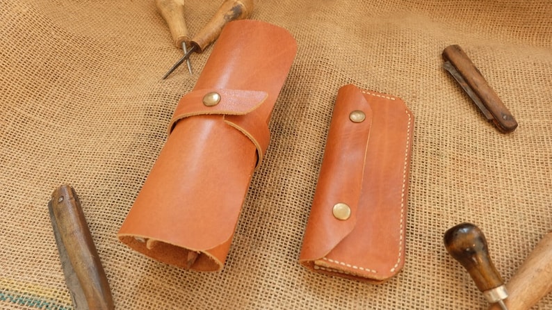 Puede incluir: Dos rollos de herramientas de cuero marr&oacute;n y un peque&ntilde;o estuche de cuero con cierres de bot&oacute;n de lat&oacute;n. Los rollos de herramientas son cil&iacute;ndricos y rectangulares, con costuras visibles. Varias herramientas de carpinter&iacute;a est&aacute;n dispersas.