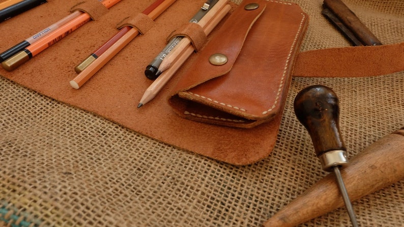 Puede incluir: Un estuche enrollable de cuero marr&oacute;n con l&aacute;pices y un bol&iacute;grafo en su interior. El estuche est&aacute; abierto, revelando los l&aacute;pices. Tambi&eacute;n se ve una peque&ntilde;a bolsa de cuero con cierre de bot&oacute;n a presi&oacute;n. Una lezna de cuero y otras herramientas est&aacute;n en el fondo.