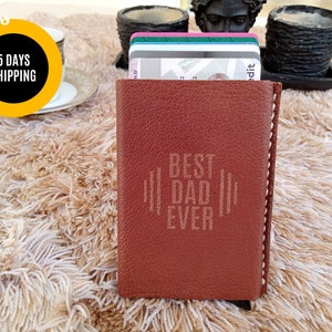 May include: A brown leather wallet with the text "BEST DAD EVER" engraved on the front. The wallet is filled with several credit cards. The background is a fluffy, light-colored rug.
