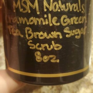 May include: A black jar with a gold lid and a handwritten label that reads "MSM Naturals Chamomile Green Tea Brown Sugar Scrub 8 oz".
