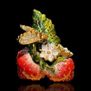 Op de afbeelding: Een gedetailleerde emaille broche met twee rode aardbeien met groene bladeren en een goudkleurige bij versierd met strass-steentjes. Er is ook een witte bloem met strass-accenten. De broche heeft een goudkleurige rand op een zwarte achtergrond.