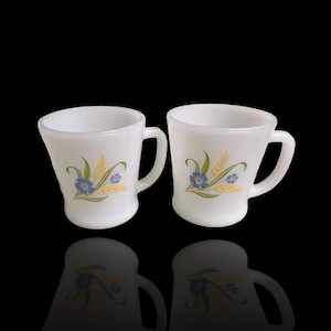 Pode incluir: Duas canecas de cerâmica branca com alças curvas. Cada caneca apresenta um design floral com flores azuis, folhas verdes e espigas de trigo amarelas. As canecas estão sobre um fundo preto com uma superfície refletora.