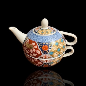 Op de afbeelding: Een decoratieve keramische theepot en kopjes set. De theepot is rond met een witte tuit, handvat en deksel. De set heeft een kleurrijk bloemen- en geometrisch patroon in rood, blauw, goud en oranje. Het kopje is aan de theepot bevestigd.