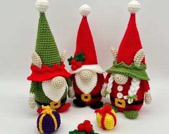 Pode incluir: Três gnomos de crochê com chapéus vermelhos e verdes e barbas brancas. Os gnomos estão segurando pequenos presentes de crochê.