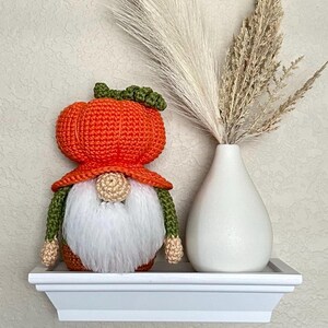 A crochet gnome featuring a large orange pumpkin-shaped hat with a green stem top. The gnome has a round beige nose, white fluffy beard, and a dark green body with a brown base.