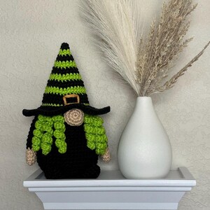 A crocheted Halloween witch gnome wearing a tall, pointed hat with horizontal black and lime green stripes, a wide black brim, a tan nose, and curly lime green hair, positioned next to a white vase filled with dried pampas grass. This is a downloadable crochet pattern PDF.