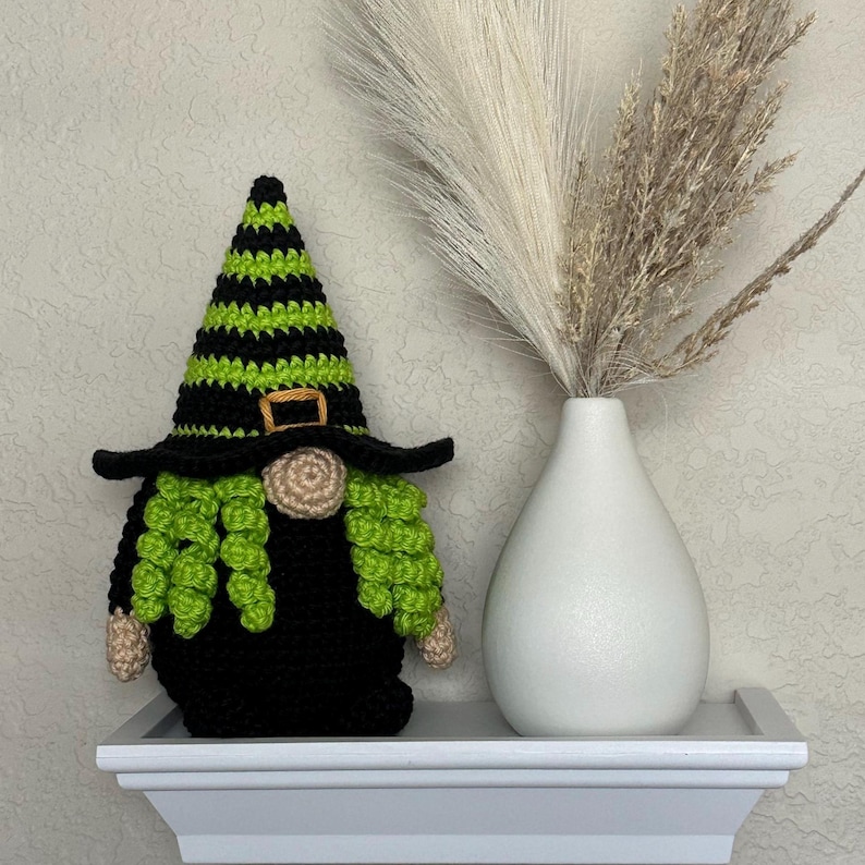 A crocheted Halloween witch gnome wearing a tall, pointed hat with horizontal black and lime green stripes, a wide black brim, a tan nose, and curly lime green hair, positioned next to a white vase filled with dried pampas grass. This is a downloadable crochet pattern PDF.