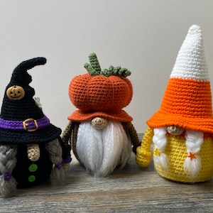 May include: Three crocheted gnomes, one dressed as a witch, one as a pumpkin, and one as a candy corn. The witch gnome has a black hat with a purple band and a buckle, a black dress, and gray braids. The pumpkin gnome has an orange pumpkin hat and a white beard. The candy corn gnome has a yellow and orange candy corn hat and a white beard.
