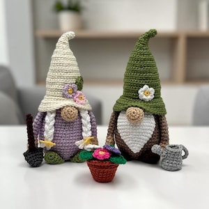 Puede incluir: Dos gnomos de jardín de ganchillo con sombreros puntiagudos, uno crema y otro verde. El gnomo crema tiene un cuerpo morado y cabello trenzado. El gnomo verde tiene un cuerpo marrón y barba blanca. Una maceta pequeña y una regadera están al frente.