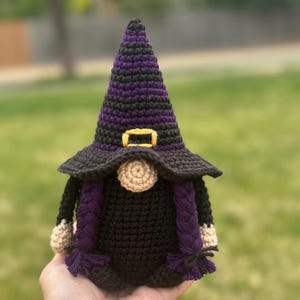 A crocheted Halloween witch gnome figurine wearing a tall, pointed purple and black hat with a yellow buckle. The gnome has a round beige nose, a black body, and two long purple braided pigtails hanging down the sides.