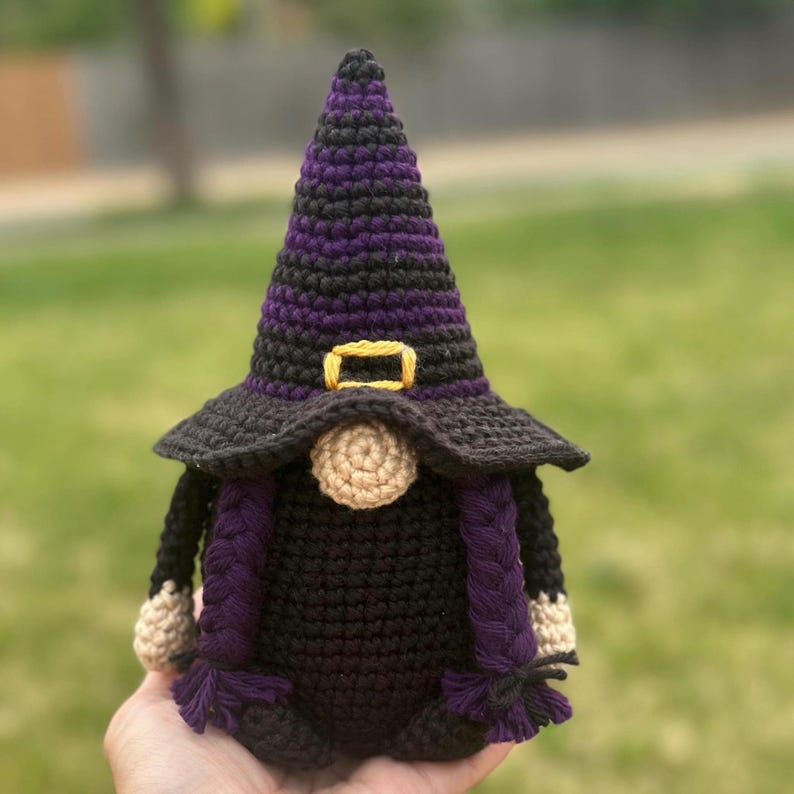 A crocheted Halloween witch gnome figurine wearing a tall, pointed purple and black hat with a yellow buckle. The gnome has a round beige nose, a black body, and two long purple braided pigtails hanging down the sides.