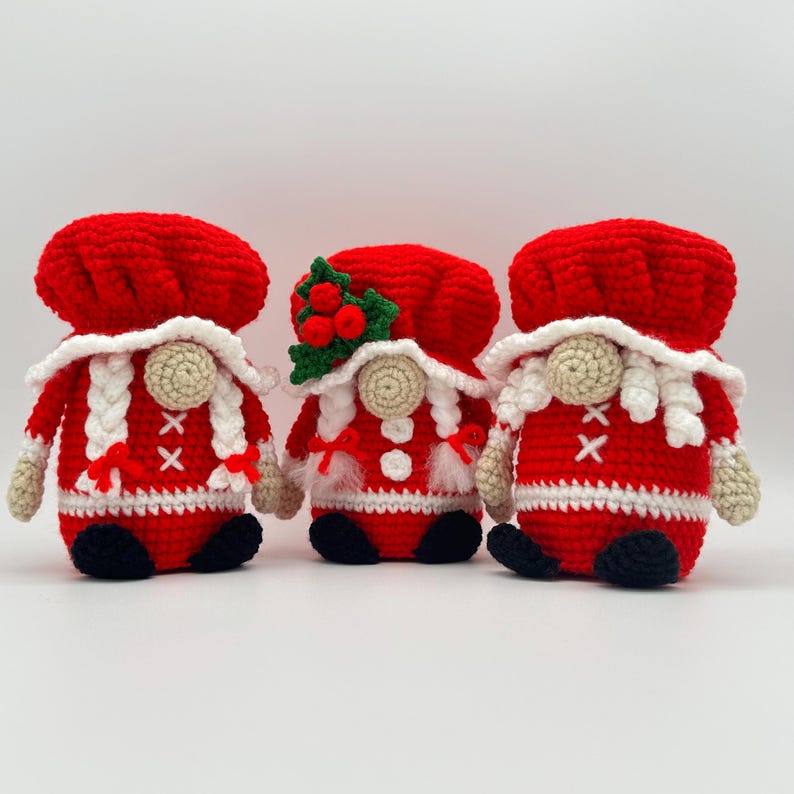 Three crochet Mrs. Claus gnomes in red and white Christmas outfits with hats, braids, and festive holly details, handmade amigurumi figures displayed for the downloadable PDF crochet pattern.