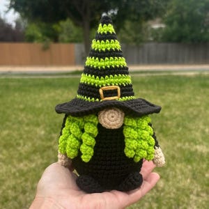 A crochet gnome figure featuring a tall, pointed hat with horizontal stripes of black and lime green yarn. The hat includes a small, rectangular yellow buckle. The gnome has a beige nose and crocheted curly lime green hair.