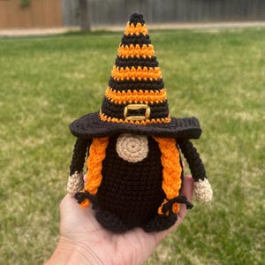 A crocheted Halloween witch gnome wearing a tall, pointed witch hat with black and orange horizontal stripes and a yellow buckle. The gnome has a round beige nose, two long orange braided pigtails, and a solid black body.