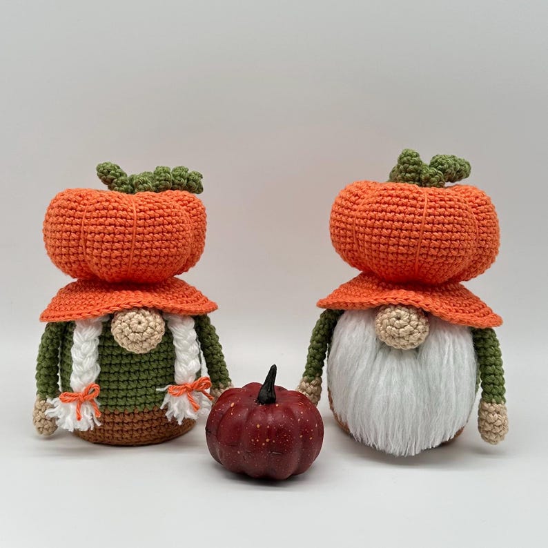 Two crocheted gnomes with orange pumpkin-shaped hats, one with white braided hair and the other with a white beard, positioned on either side of a small, dark red, textured decorative pumpkin figurine. Digital PDF download.