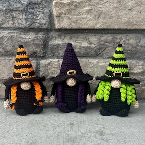 Three crocheted witch gnomes wearing pointed hats with gold-colored buckles. The left gnome has orange and black stripes with orange braids, the center has a purple and black hat with purple braids, and the right has lime green and black stripes. This is a downloadable PDF crochet pattern.