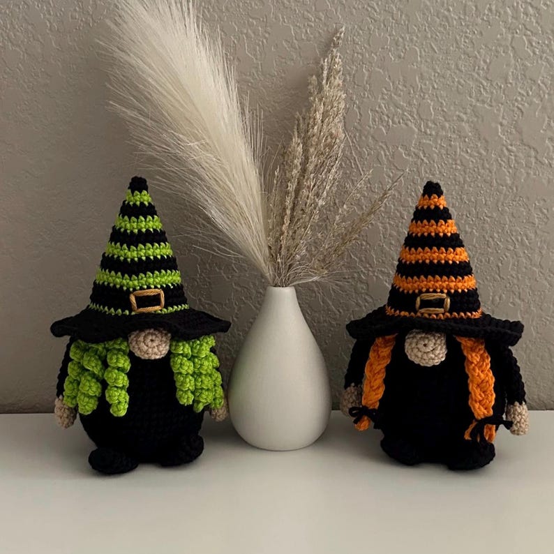 Two crocheted witch gnomes wearing black pointed hats with horizontal stripes in lime green and orange respectively. Each hat features a small gold-colored buckle. The gnomes have round beige noses, black bodies, and long hair matching their hat colors.