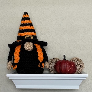 A crocheted witch gnome wearing a tall, black and orange striped pointed hat and a wide-brimmed black hat. The gnome has a beige nose, two long orange braided pigtails, and a solid black body. Beside it sits a small red pumpkin.