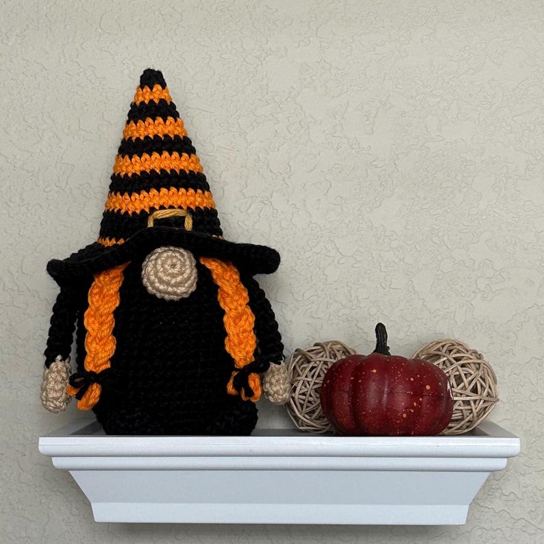 A crocheted witch gnome wearing a tall, black and orange striped pointed hat and a wide-brimmed black hat. The gnome has a beige nose, two long orange braided pigtails, and a solid black body. Beside it sits a small red pumpkin.
