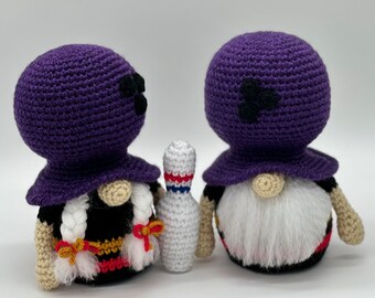 May include: Two purple crocheted gnomes with white beards and black hats. The gnomes are standing next to a white crocheted bowling pin.