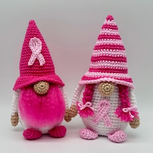 Puede incluir: Dos gnomos de ganchillo hechos a mano. El gnomo de la izquierda tiene un sombrero de color rosa intenso con una aplicación de lazo rosa claro y una barba de piel sintética rosa brillante. El gnomo de la derecha tiene un sombrero a rayas rosas y blancas, dos trenzas rosas y un cuerpo blanco con una aplicación de lazo rosa claro.