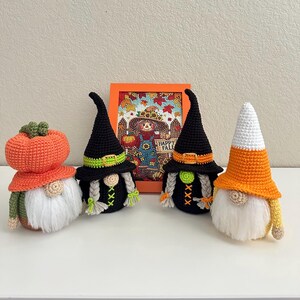 May include: Four crocheted gnomes with white yarn beards and round noses. One has an orange pumpkin hat, two wear black witch hats with green or orange bands and braided hair, and one has a white and orange candy corn hat. A framed picture sits behind.