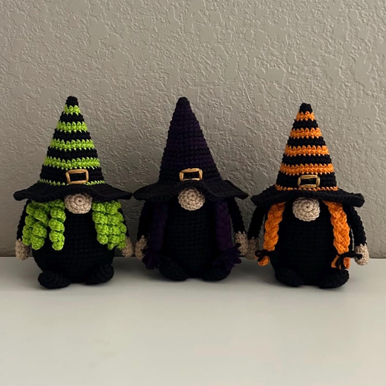 Three crocheted witch gnomes with black bodies and pointed hats. The first has lime green stripes and hair, the second has purple stripes and hair, and the third has orange stripes and hair. Each hat features a gold-colored buckle detail.