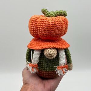 A crochet gnome featuring a large orange pumpkin-shaped hat with a green stem top. The gnome has a round beige nose, two white braided yarn pigtails tied with small orange bows, and a dark green body with a brown base.