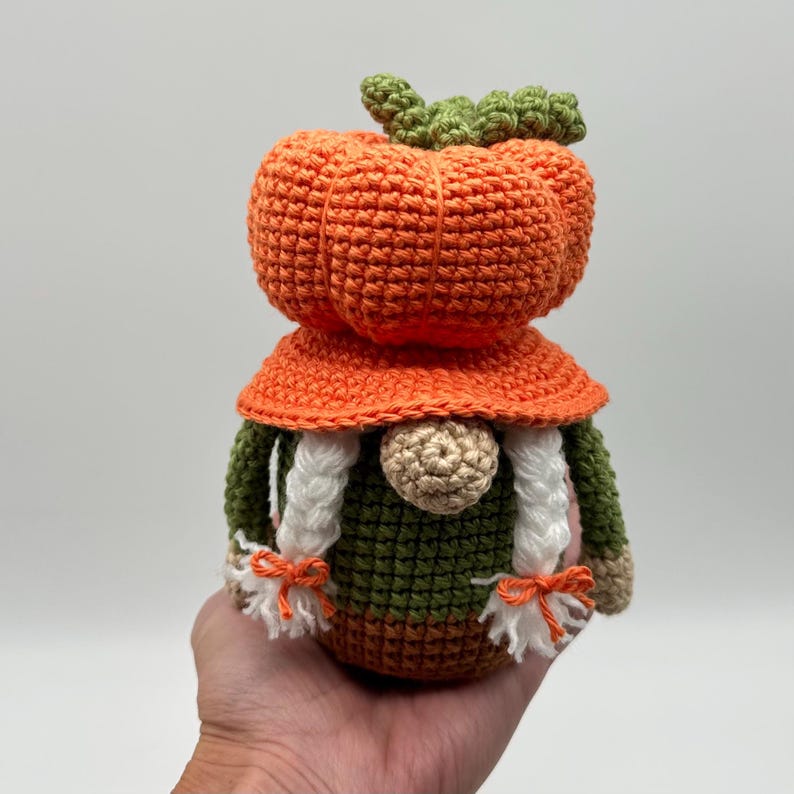 A crochet gnome featuring a large orange pumpkin-shaped hat with a green stem top. The gnome has a round beige nose, two white braided yarn pigtails tied with small orange bows, and a dark green body with a brown base.